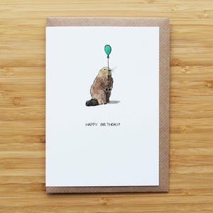 May include: A birthday card with a watercolour illustration of a beaver holding a teal balloon. The beaver is brown and grey, with a black tail. The words "HAPPY BIRTHDAY!" are printed below the beaver.