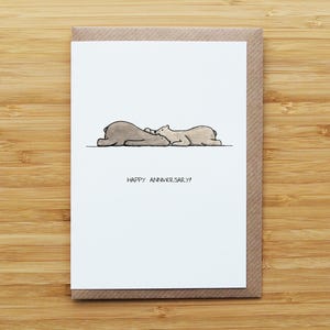 May include: A greetings card with a brown envelope. The white card shows a minimalist illustration of two bear-like figures lying down, with the text "HAPPY ANNIVERSARY!" beneath the illustration.