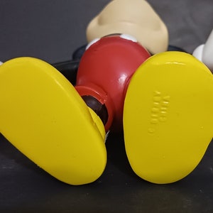 K&ouml;nnte beinhalten: Eine gelbe Plastikfigur von Mickey Mouse mit roten Shorts und schwarzen Schuhen. Die Schuhe haben den Text "Disney" auf der Unterseite.