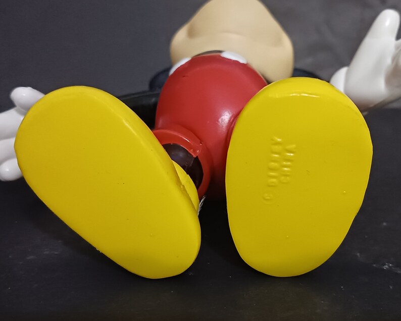 K&ouml;nnte beinhalten: Eine gelbe Plastikfigur von Mickey Mouse mit roten Shorts und schwarzen Schuhen. Die Schuhe haben den Text "Disney" auf der Unterseite.