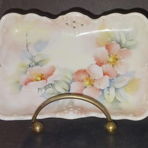 May include: A white porcelain serving dish with a delicate floral design featuring pink roses and green leaves. The dish has a scalloped edge and a pierced design around the rim.