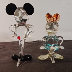 May include: Two glass figurines, one resembling Mickey Mouse with black ears and a red button, the other a female character with a brown headpiece and blue accents. Both are transparent and stand on a dark surface.