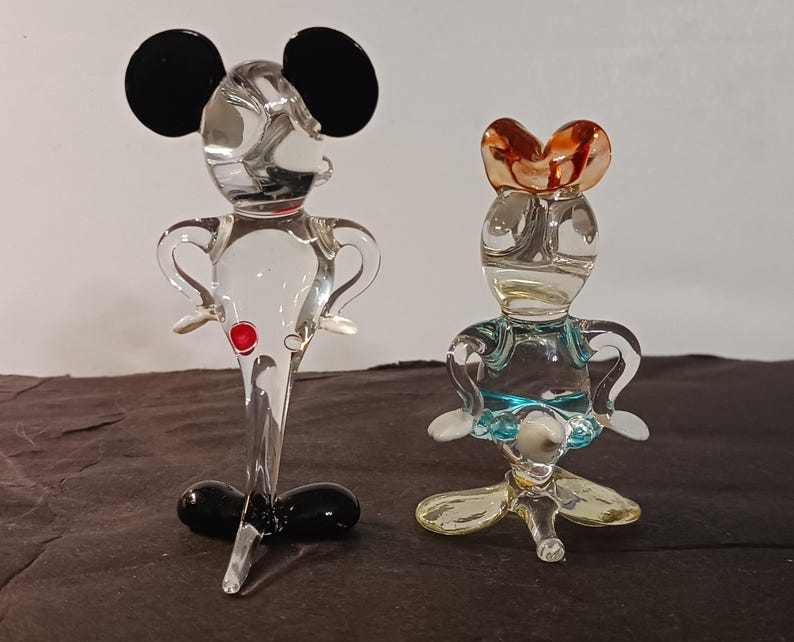 May include: Two glass figurines, one resembling Mickey Mouse with black ears and a red button, the other a female character with a brown headpiece and blue accents. Both are transparent and stand on a dark surface.