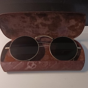 May include: Round sunglasses with dark lenses and a thin metallic frame resting inside an open, weathered brown hard-shell case. The interior of the case features a textured, worn lining, while the exterior shows signs of age and surface wear.