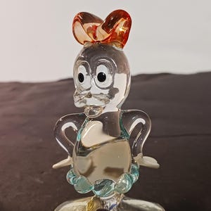 May include: A clear glass figurine of a duck with a bow on its head. The duck has large black eyes, a beak, and a blue collar. The bow is a reddish-orange color.