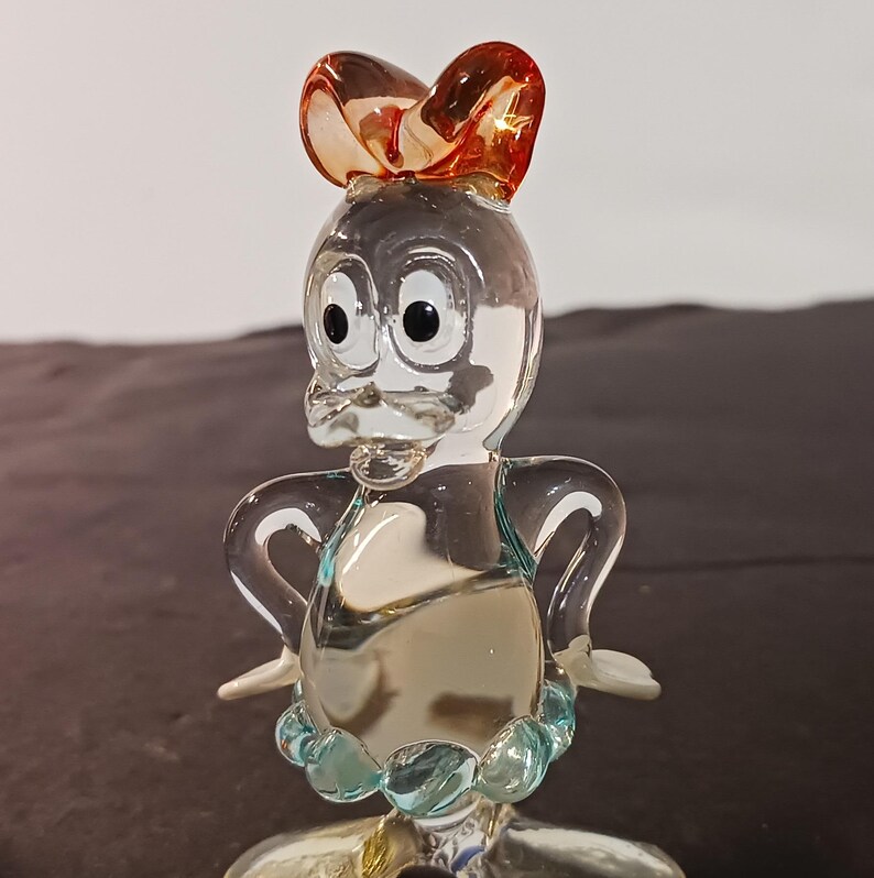 May include: A clear glass figurine of a duck with a bow on its head. The duck has large black eyes, a beak, and a blue collar. The bow is a reddish-orange color.