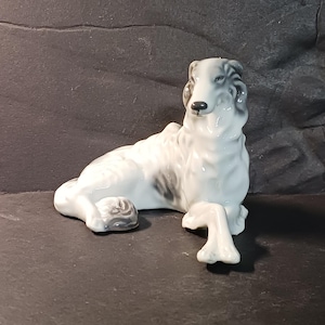 May include: A porcelain figurine of a Borzoi dog in a reclining position. The dog is primarily white with grey shading on its ears, back, and tail. It has a long, slender snout, dark eyes, and a black nose, with one front leg extended forward.