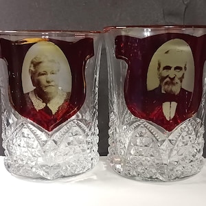May include: Two clear glass tumblers featuring a cut crystal diamond pattern on the lower half. Each glass has a deep red decorative shield shape near the rim, containing a sepia-toned oval portrait of an elderly person in formal attire.