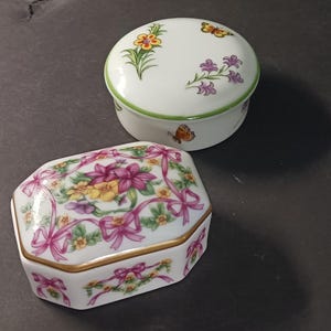 May include: Two decorative porcelain trinket boxes with floral and butterfly designs. The larger box is octagonal and features pink bows and flowers. The smaller box is round and features a green trim and a butterfly.