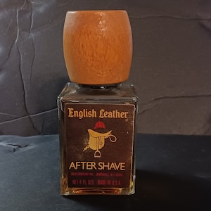 May include: A brown glass bottle of English Leather After Shave. The bottle has a wooden cap and a label with a saddle and a riding crop. The label also says "English Leather" and "After Shave". The bottle is 4 fl oz and made in the USA.
