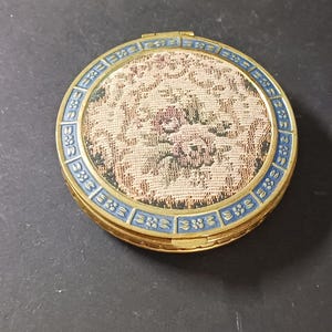 May include: A gold compact with a blue and gold floral design. The compact has a hinged lid and a mirror inside.