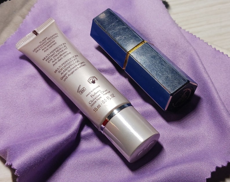 May include: A silver tube with a blue cap and a white label with black text. The text on the label reads "Flash Defatigant Regard Multi-Perfection Eye Treatment Anti-poches Multi-Perfection Instant Rescue Eye Treatment Anti-puffiness Anti-dark circles Multi Perfektion Soforteffekte Augenpflege Gegen Schwellungen und Augenschatten GM Made in France Parfums Christian Dior 75000 Paris 15 ml - 0.5 fl oz". A blue and gold lipstick tube is also in the image.