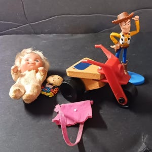 May include: A set of vintage toys including a doll, a toy motorcycle, a doll, and a pink purse. The doll is wearing a cream-colored outfit and has blonde hair. The motorcycle is red and has black wheels. The doll is wearing a colorful outfit. The purse is made of pink fabric.