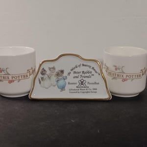 May include: A set of Beatrix Potter themed porcelain items. Includes two white cups with "Beatrix Potter" text and floral accents, and a decorative tray featuring Peter Rabbit and friends. The tray has a gold trim and text.