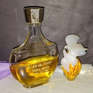 May include: Two bottles of L'Air du Temps perfume by Nina Ricci. The larger bottle is clear glass with a gold cap and the smaller bottle is a gold-colored glass with a white plastic cap. Both bottles contain a yellow liquid.