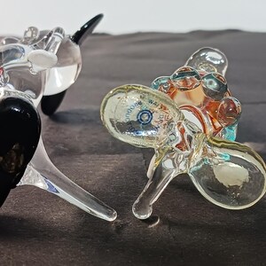 May include: Two glass sculptures, one resembling a whale with black and white accents, the other a cluster of clear orbs with a colorful interior. The sculptures are on a dark surface.
