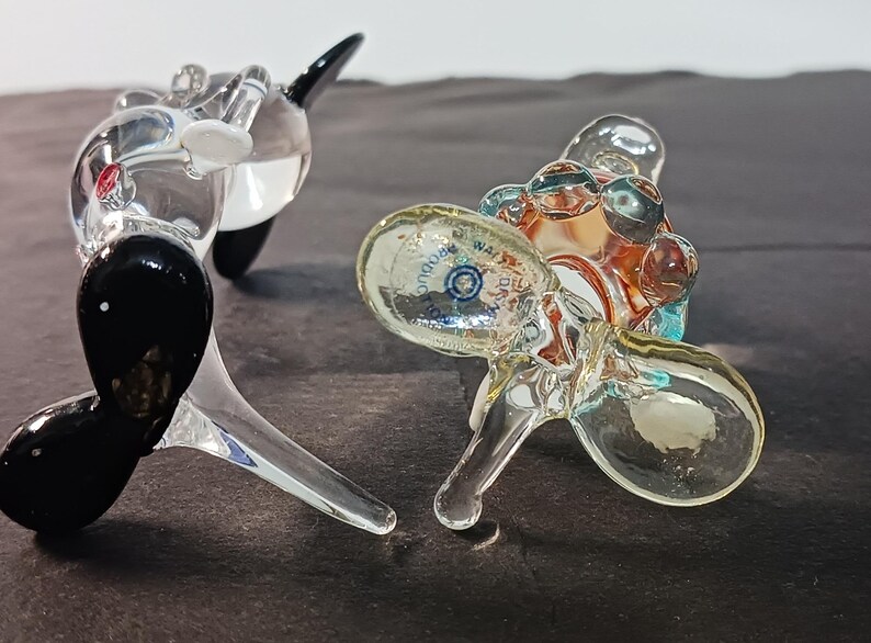 May include: Two glass sculptures, one resembling a whale with black and white accents, the other a cluster of clear orbs with a colorful interior. The sculptures are on a dark surface.