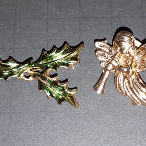 May include: Two gold-tone Christmas ornaments. One is a holly sprig with green enamel leaves. The other is a gold angel with wings and a trumpet.