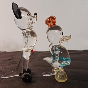 May include: Two clear glass figurines of cartoon characters. One is a mouse with black ears and a red button. The other is a duck wearing a hat and a blue skirt. Both figurines have black feet and are standing on a dark surface.