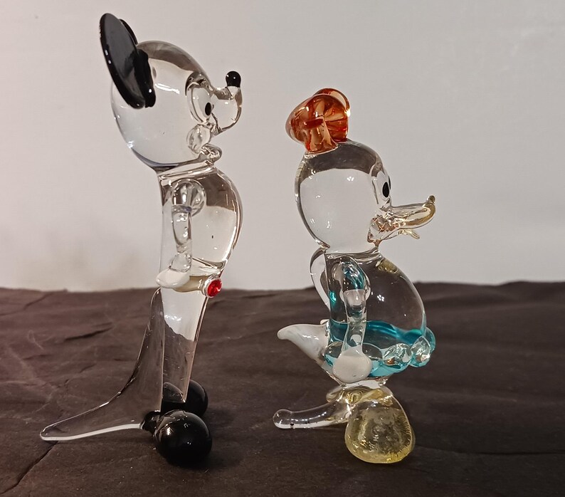May include: Two clear glass figurines of cartoon characters. One is a mouse with black ears and a red button. The other is a duck wearing a hat and a blue skirt. Both figurines have black feet and are standing on a dark surface.