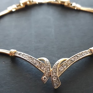 May include: A gold chain necklace with a delicate, crystal-encrusted pendant. The pendant features a double-loop design with a V-shaped silhouette.