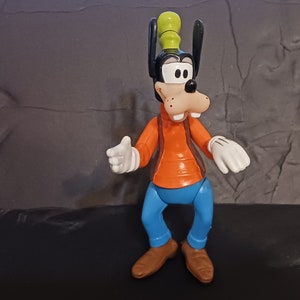 K&ouml;nnte beinhalten: Eine Plastikfigur von Goofy, einer Zeichentrickfigur von Disney. Goofy tr&auml;gt eine braune Weste, ein orangefarbenes Hemd, eine blaue Hose und einen gr&uuml;nen Hut. Er hat ein breites L&auml;cheln und h&auml;lt seine H&auml;nde vor sich.