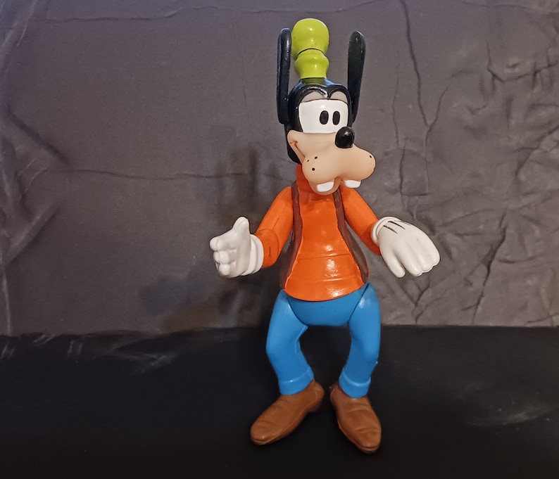 K&ouml;nnte beinhalten: Eine Plastikfigur von Goofy, einer Zeichentrickfigur von Disney. Goofy tr&auml;gt eine braune Weste, ein orangefarbenes Hemd, eine blaue Hose und einen gr&uuml;nen Hut. Er hat ein breites L&auml;cheln und h&auml;lt seine H&auml;nde vor sich.
