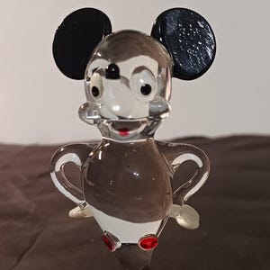 May include: A clear glass figurine of a cartoon mouse with black ears, a white face, and a brown body. The mouse has red accents on its feet and mouth. The figurine is standing with its arms out.
