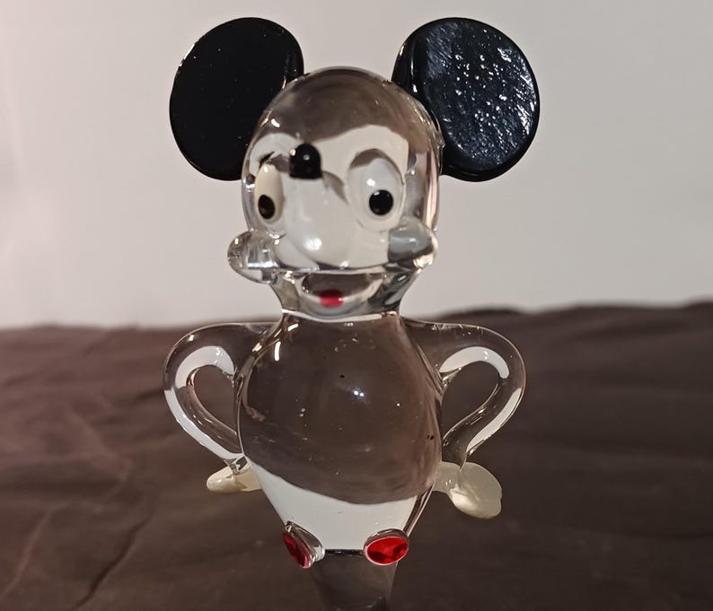 May include: A clear glass figurine of a cartoon mouse with black ears, a white face, and a brown body. The mouse has red accents on its feet and mouth. The figurine is standing with its arms out.