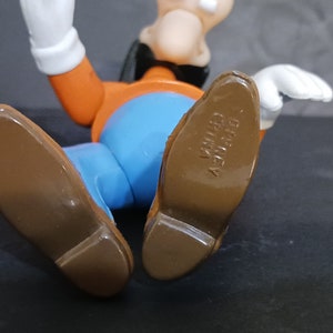 K&ouml;nnte beinhalten: Eine braune und orangefarbene Comicfigur mit einem blauen Hemd und braunen Schuhen. Die Schuhe haben den Text "DISNEY" und "CHINA" auf der Unterseite.