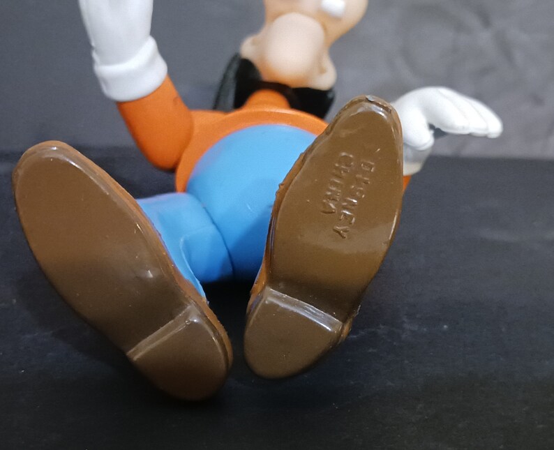 K&ouml;nnte beinhalten: Eine braune und orangefarbene Comicfigur mit einem blauen Hemd und braunen Schuhen. Die Schuhe haben den Text "DISNEY" und "CHINA" auf der Unterseite.