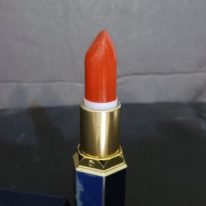 May include: A red lipstick in a gold and blue tube. The lipstick is in a bullet shape and is ready to be applied.