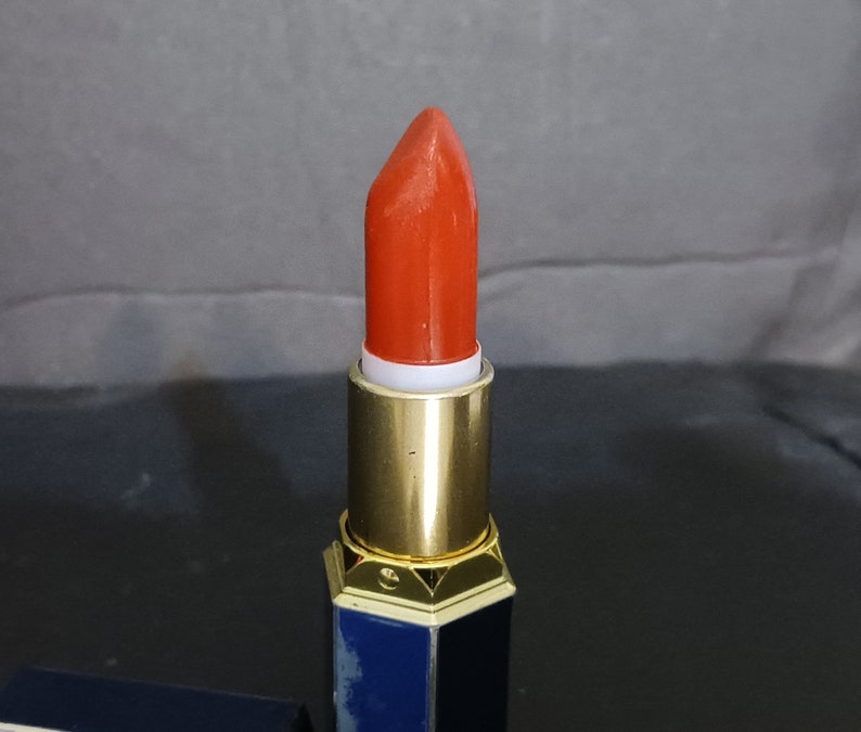 May include: A red lipstick in a gold and blue tube. The lipstick is in a bullet shape and is ready to be applied.