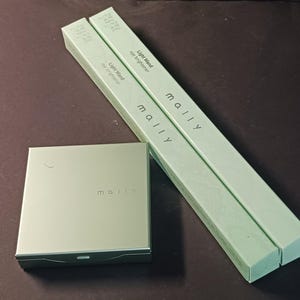 May include: Two green and white cardboard boxes with the word "Mally" printed on them. The boxes are stacked on top of each other. The top box has the text "Light Wand Eye Brightener" printed on it. The bottom box has the text "Light Wand Eye Brightener" printed on it. A small green and white box with the word "Mally" printed on it is in the foreground.