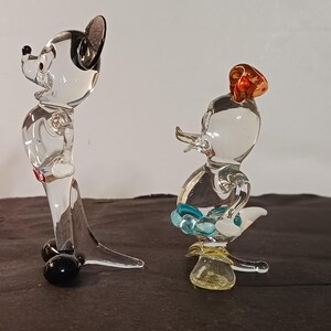 May include: Two clear glass figurines, one resembling a mouse with black ears and a red accent, and the other a duck with a red hat and blue accents. The figurines are detailed and stand on a dark surface.
