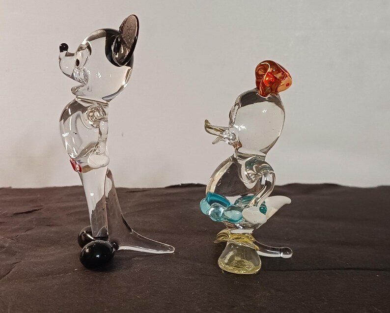 May include: Two clear glass figurines, one resembling a mouse with black ears and a red accent, and the other a duck with a red hat and blue accents. The figurines are detailed and stand on a dark surface.