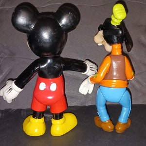 K&ouml;nnte beinhalten: Eine schwarze und rote Mickey Mouse Figur und eine braune, orange und blaue Goofy Figur. Beide Figuren stehen auf einer grauen Oberfl&auml;che.