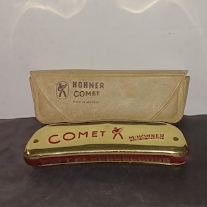 May include: A gold and red Hohner Comet harmonica with its tan case. The harmonica has the word "COMET" in red letters and the Hohner logo. The case has the Hohner logo and the words "Hohner Comet Made in Germany".