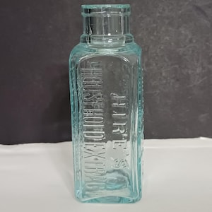 May include: A vintage, rectangular, light blue glass bottle with embossed text reading "HOUSEHOLD EXTRACTS". The bottle has a square shape with rounded edges and a short neck. The bottle is empty and sits on a white surface.