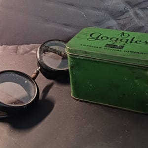 May include: A green metal box with the text "AO Goggles Made in USA American Optical Company" on the lid. The box is next to a pair of black goggles with round lenses.