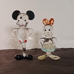 May include: Two clear glass figurines of cartoon characters. One is a mouse with black ears, red accents, and a black base. The other is a duck with an orange bow, blue and yellow accents. Both have outstretched arms.