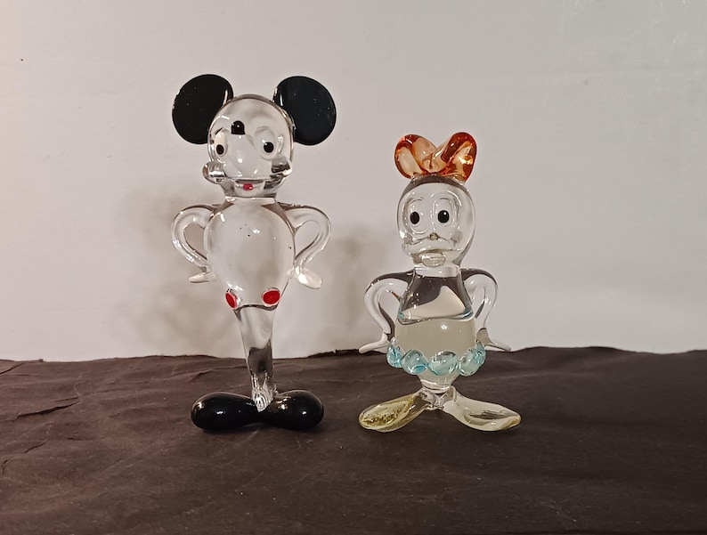 May include: Two clear glass figurines of cartoon characters. One is a mouse with black ears, red accents, and a black base. The other is a duck with an orange bow, blue and yellow accents. Both have outstretched arms.