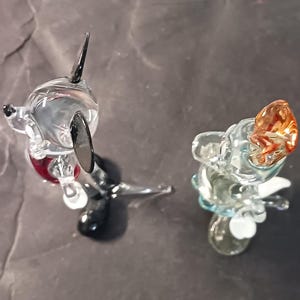 May include: Two glass figurines, one resembling a dog with black ears and a red collar, and the other a small creature with an orange head. Both are transparent and sit on a dark surface.