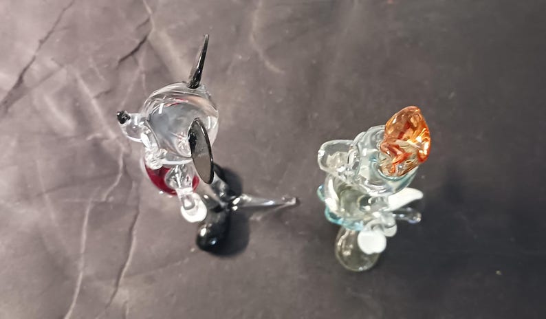 May include: Two glass figurines, one resembling a dog with black ears and a red collar, and the other a small creature with an orange head. Both are transparent and sit on a dark surface.