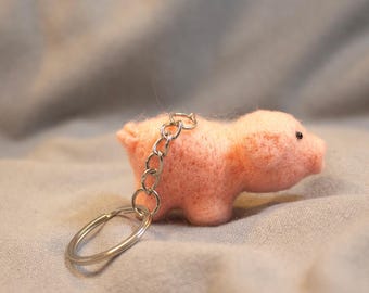May include: A small, pink felt pig keychain with a silver chain and keyring. The pig has tiny black eyes and a short tail. The keychain is set against a soft, gray background.