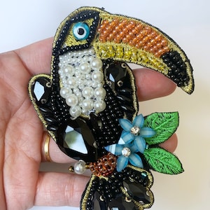 May include: A toucan brooch made of black, white, yellow, orange, and blue beads. The toucan's beak is made of orange and yellow beads, and its eye is a blue and white bead. The toucan is surrounded by green leaves and blue flowers with rhinestones.