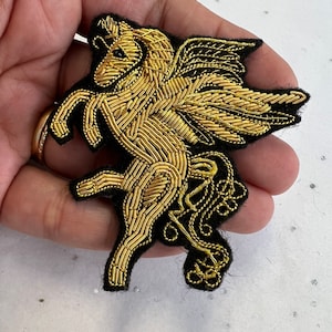 May include: A gold embroidered winged horse patch with black felt backing. The horse is in a rearing pose with its wings spread out.
