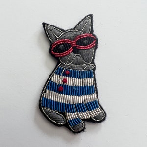 May include: Embroidered patch of a gray dog wearing red sunglasses and a blue and white striped sweater. The dog has a black outline and is sitting upright. The sweater has three red buttons.