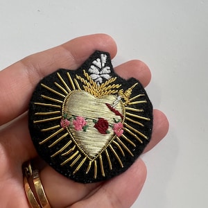 May include: A gold heart with a red and pink rose design, surrounded by gold rays, embroidered on black felt.