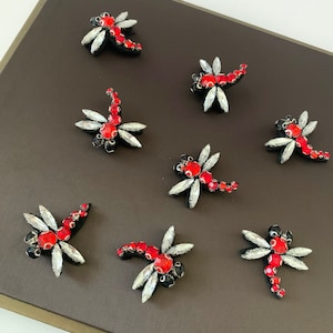 Peut inclure: Huit embellissements de libellule rouge et argent sur une surface brun foncé. Les libellules sont faites de strass et ont des accents noirs.